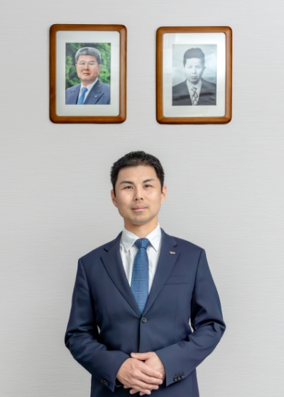 初代社長と二代目社長の写真と並んで立つ代表取締役